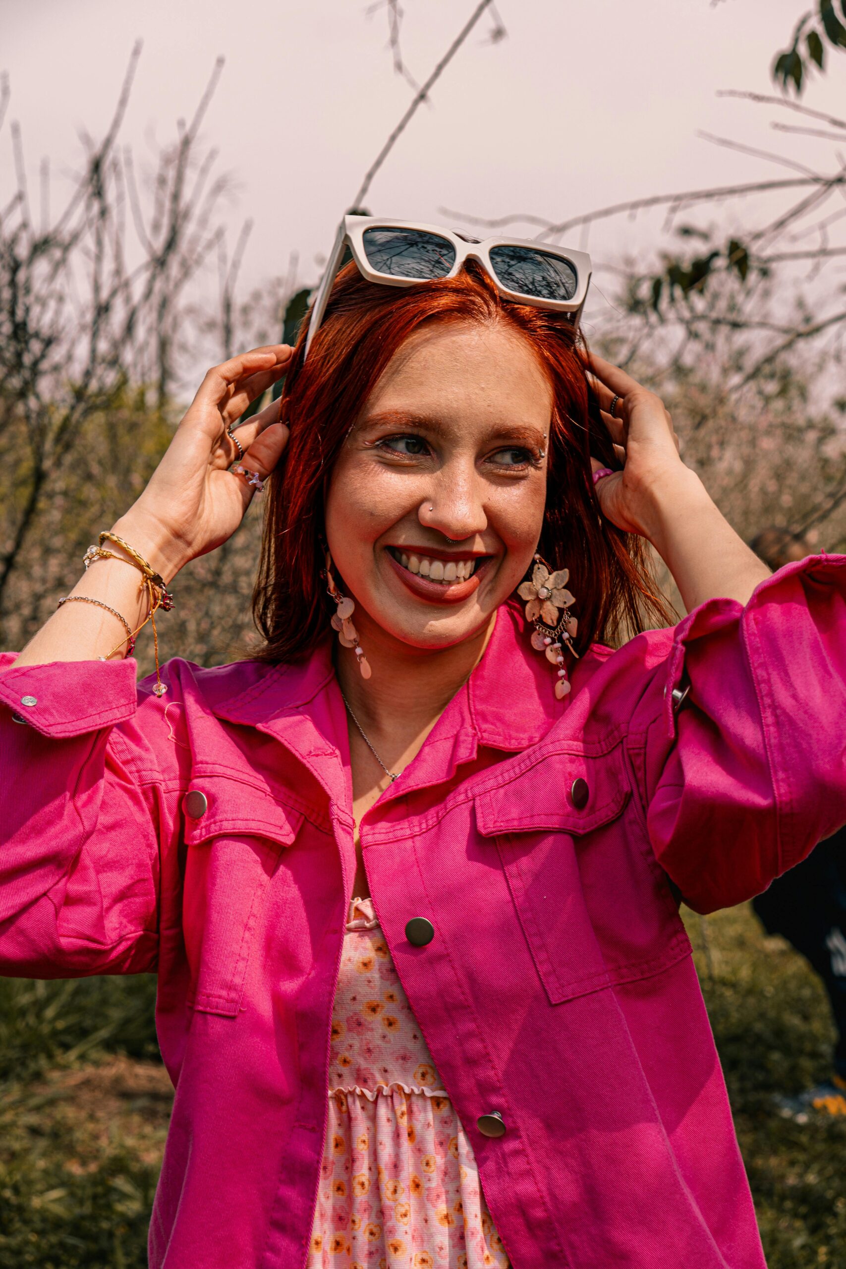 Smiling woman in a vibrant pink jacket enjoying a sunny day outdoors with stylish accessories.